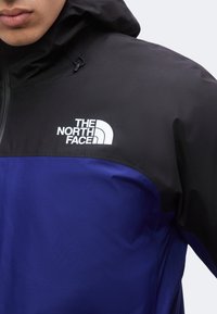 Άτομο που φοράει ένα μαύρο και μπλε μπουφάν North Face με κουκούλα, με το λογότυπο της μάρκας στο πάνω μέρος του στήθους.