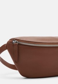 Borsa in pelle marrone con superficie strutturata, zip in argento e manico di trasporto piccolo. Etichetta impressa su un lato. Design compatto e arrotondato.