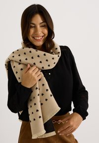 Écharpe beige avec des motifs de cœurs noirs, fabriquée en matériau doux, avec des bords frangés, enroulée autour des épaules d'une personne.