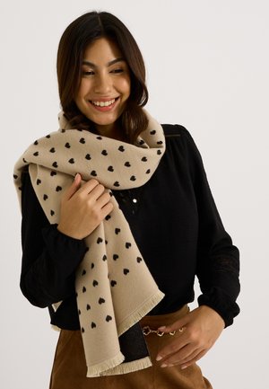 Bufanda beige con patrones de corazones negros, hecha de material suave, con bordes con flecos, enrollada alrededor de los hombros de una persona.