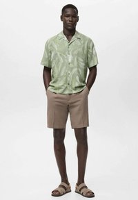 Camisa de manga corta verde con patrón abstracto, combinada con pantalones cortos beige y sandalias marrones. Tejido suave, corte relajado. Sin logotipos visibles.