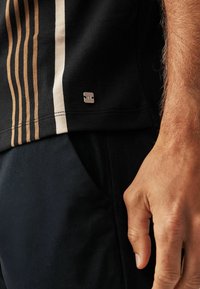 T-shirt noir avec des rayures beige, texture lisse et un petit logo métallique sur l'ourlet, associé à un pantalon de couleur foncée.