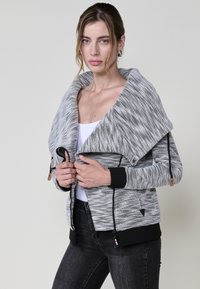 Szürke és fehér mintás dzseki, nagy sállal, cipzáras záródással, fekete díszítéssel a mandzsettákon és az alján. Deréknál húzózsinórral rendelkezik.