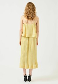 Vestido amarillo plisado con tirantes ajustables, diseño en capas, tela fluida y dobladillo a la altura de la rodilla, combinado con zapatos negros de tacón alto.