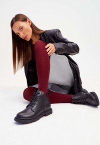 Botine din piele neagră, până la gleznă, cu sistem de șiret, vârf rotund și talpă texturată. Modelul poartă colanți bordeaux și un blazer elegant negru.