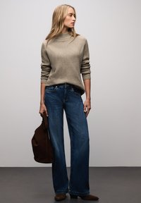 Pull beige côtelé à col montant, associé à un jean en denim bleu évasé. Le mannequin porte un sac en daim marron foncé.