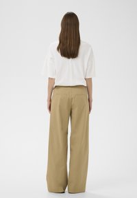 Femme aux longs cheveux bruns se tenant de dos, portant un haut blanc à manches courtes et amples et un pantalon beige à jambes larges.