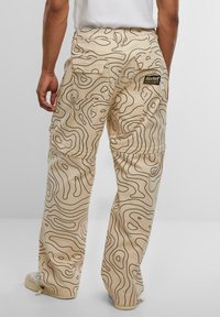 Beige cargo pants with a wavy black pattern, featuring back pockets and a logo patch. Made of fabric with a smooth texture.