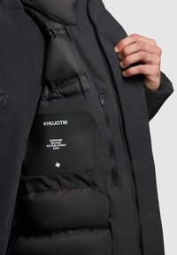 Czarna kurtka z gładkiego materiału zewnętrznego, puchowym wnętrzem i kieszenią na zamek z etykietą marki i logo.