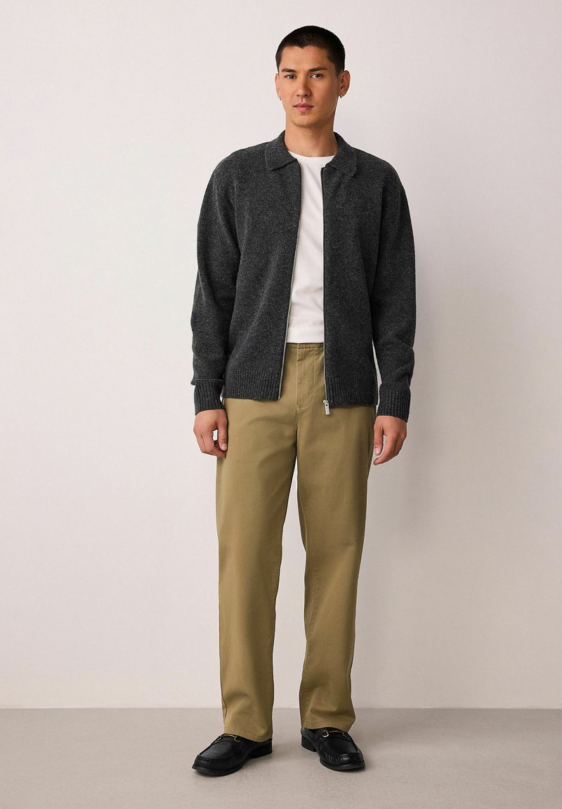 Šedý ležérny cardigan na zips s golierom, rebrovanými manžetami a voľným strihom, kombinovaný so svetlými khaki nohavicami a čiernymi topánkami. Hladká textura.