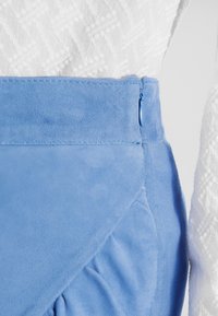 Jupe en daim bleu clair avec une surface texturée, présentant un design angulaire et une fermeture éclair latérale dissimulée. Haut blanc à motifs partiellement visible.