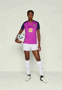 Jucătoare de fotbal îmbrăcată în uniformă de culoare mov și albă, ținând o minge de fotbal și stând pe un fundal simplu, pregătită să joace.