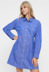 Frau, die ein blau-weiß gestreiftes, knöpfbares Hemdkleid mit langen Ärmeln trägt, steht mit einer Hand an der Hüfte und schaut seitlich.