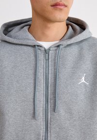 Gros plan d'une personne portant un sweat à capuche gris zippé avec cordons et un logo blanc sur la poitrine gauche.