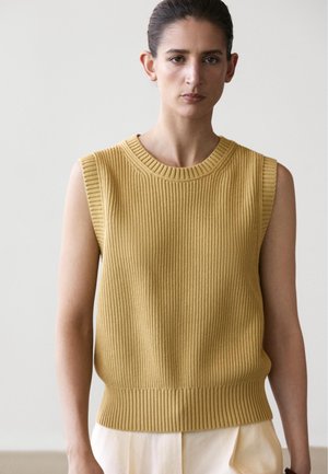 Vrouw met donker haar draagt mouwloze mosterdgele geribbelde trui en lichte broek tegen een effen achtergrond.