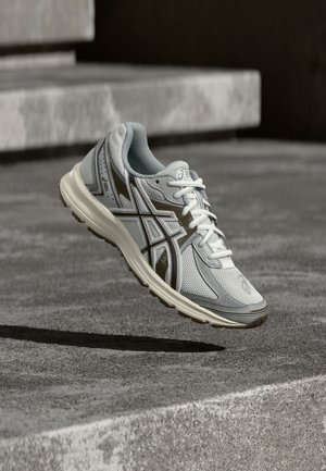 Hellegraue und weiße Asics-Laufschuhe mit braunen Akzenten, die über Betontreppen schweben und einen Schatten werfen.