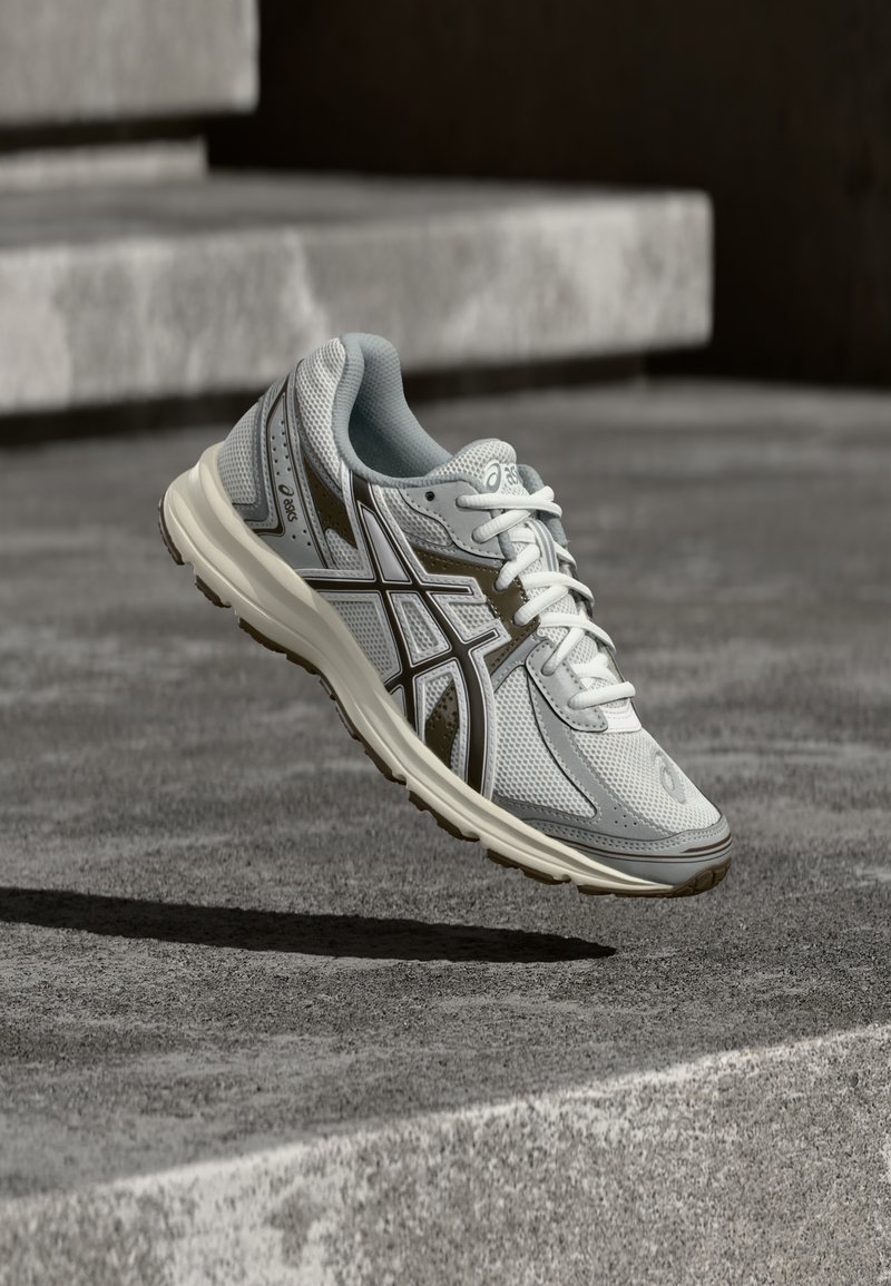 Zapatilla de running Asics en gris claro y blanco con detalles en marrón, flotando sobre unos escalones de concreto y proyectando una sombra.