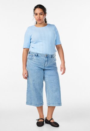 Kvinde stående iført lyseblå striktrøje med korte ærmer, højtaljede vide cropped jeans og sorte mesh Mary Jane-sko på hvid baggrund.