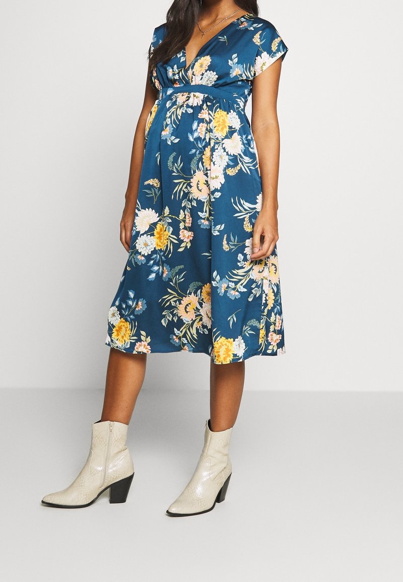 Raskausnaisen sinisessä kukkakuosisessa midi-mekossa, jossa on lyhyet hihat, ja vaaleissa keskipitkillä nilkkabootseilla, seisovan yksinkertaista taustaa vasten.