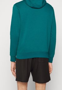 Tyrkysová mikina s kapucí a dlouhými rukávy, spárovaná s černými síťovanými šortkami s kapsou na zadní straně. Měkká textura a volný střih.