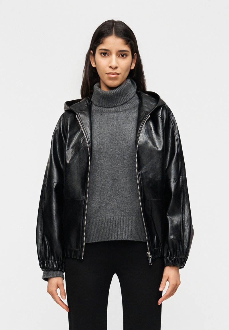 Νεαρή γυναίκα που φοράει γκρι πουλόβερ με ζιβάγκο και μαύρο δερμάτινο μπουφάν με φερμουάρ, καθώς και μαύρο παντελόνι, στέκεται μπροστά σε απλό λευκό φόντο.