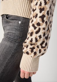 Beige gestrickter Pullover mit braunem Leopardenmuster am Ärmel, gerippte Bündchen, kombiniert mit dunkelgrauen Jeans. Strukturierte Stoffdetails sichtbar.