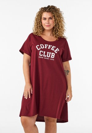Žena s kučeravými vlasmi, ktorá má na sebe voľné bordové šaty s bielym nápisom "Coffee Club, Brew, Drink, Love" na prednej strane.