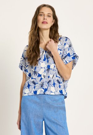 Frau mit langen braunen Haaren, die eine blau-weiß geblümte Kurzarmbluse und hellblaue Hosen trägt, posiert mit einer Hand in Brusthöhe.