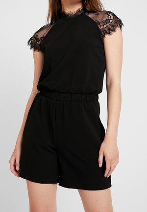 Kvinde iført en sort ærmeløs romper med blonder på skuldre og halsudskæring, stående foran en ensfarvet baggrund.