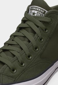 Suur lähedalt vaadatuna oliivirohelise Converse tennise taimelised paelad, metallist augud ja must-valge kummist tallatükk.