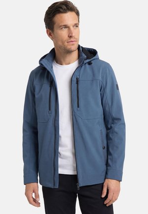 Mann trägt blaue Kapuzenjacke mit Reißverschluss über weißem Hemd, steht und blickt seitlich vor einem neutralen Hintergrund.