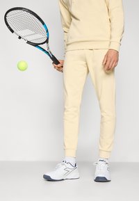 Tennisoutfit med en lys beige hættetrøje og matchende sweatpants, som holder en sort og blå tennisracket med en grøn tennisbold i luften.