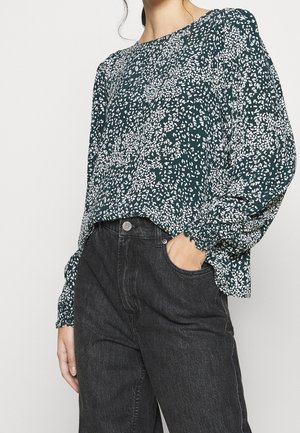 Femme portant un chemisier vert foncé à manches longues avec un motif floral blanc et un jean noir taille haute, main dans la poche.