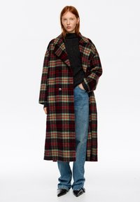 Mujer vistiendo un abrigo largo de cuadros verdes y rojos, suéter negro, jeans azules y zapatos negros puntiagudos, de pie contra un fondo blanco.