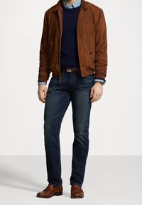 Muž oblečený v hnědé semišové bundě, námořnickém svetrě, modrých džínách, hnědném pásku, hnědých ko žených botách a s hodinkami, stojící na jednolitém pozadí.