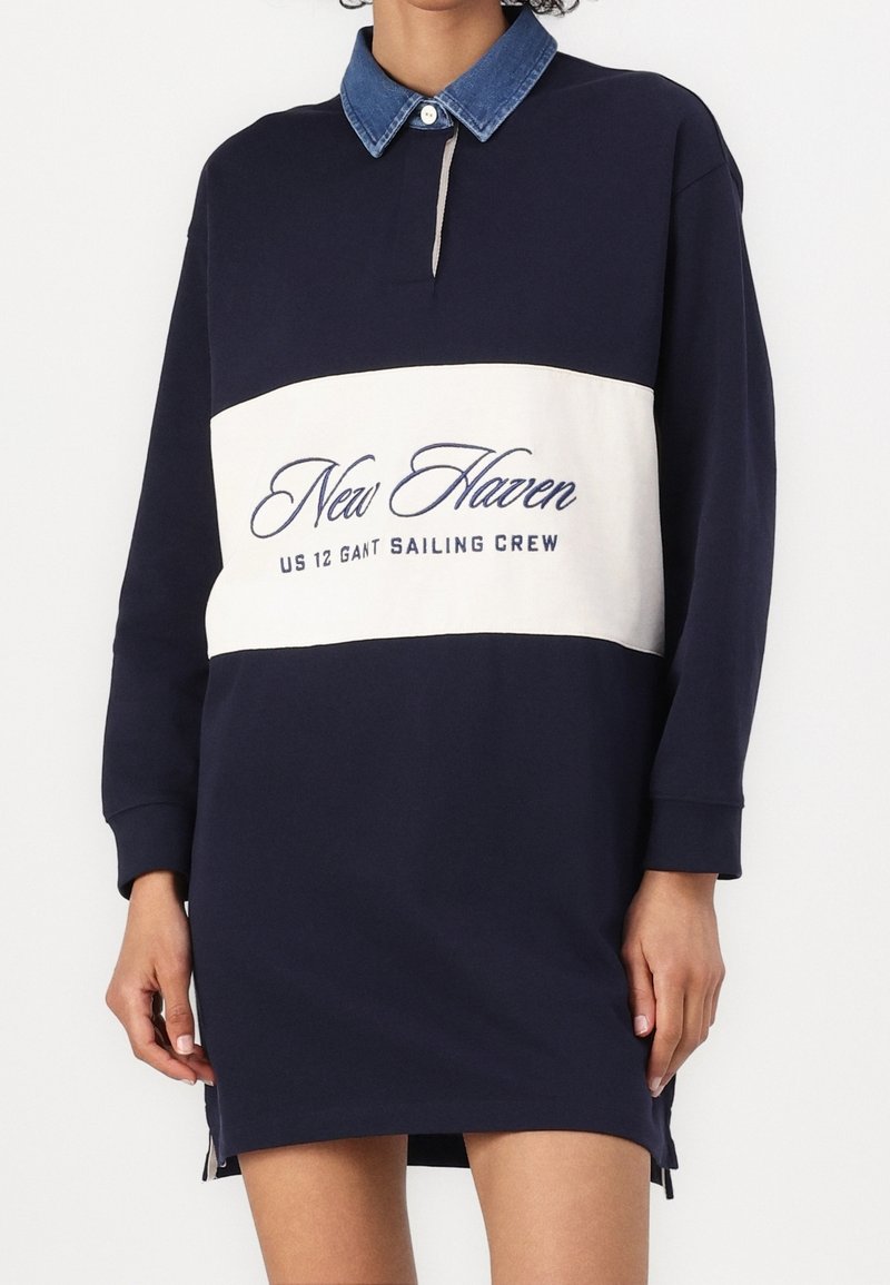 Robe bleu marine à manches longues avec panneau blanc et texte "New Haven US 12 Gant Sailing Crew", avec col en denim bleu et bouton.