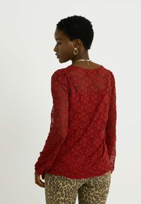 Haut en dentelle rouge à manches longues avec des motifs ondulés, au design ajusté et en tissu texturé. Accompagné d'un pantalon à imprimé léopard. Bijoux en or visibles.