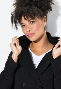 Schwarze doppelte Jacke mit großen Knöpfen, breitem Kragen und Schulterklappen, getragen über einem weißen V-Ausschnitt-Shirt. Lockige Haare sind sichtbar.