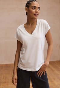 Weißes V-Ausschnitt-T-Shirt mit kurzen Ärmeln, hergestellt aus weichem Stoff. Es hat eine lockere Passform und minimale Nähte. Getragen zu einer dunklen Hose.