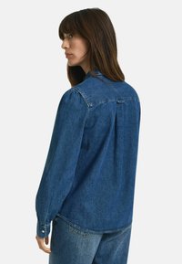Chemise en denim bleu profond avec un col à boutons, des manches longues et un détail de pli au dos. Texture douce et design décontracté.