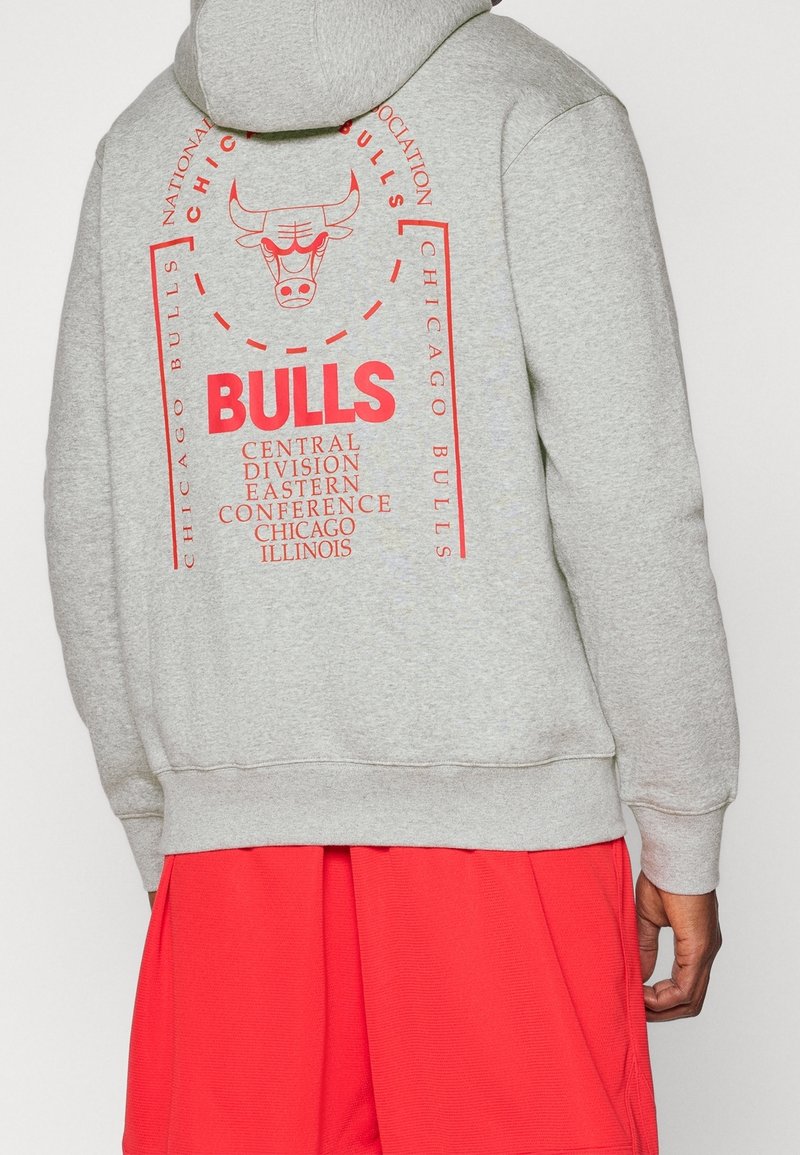 Personne portant un sweat à capuche gris avec le logo rouge des Chicago Bulls et un texte, associé à un short rouge, vue de dos.