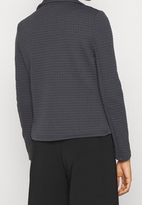 Veste matelassée grise avec une surface texturée, des manches longues et un col. L'ourlet inférieur est droit.