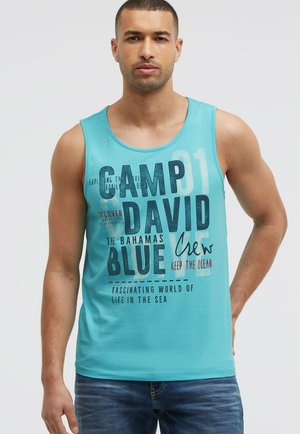 Young man in a turquoise sleeveless shirt with "Camp David Blue" and ocean-themed text, wearing blue jeans, posing against a light background.