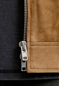 Brun ruskindsjakke panel med en sølvmetal lynlås, der viser tekstureret overflade og syningsdetaljer mod en mørk stofbaggrund.