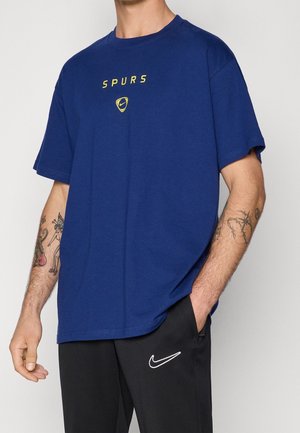 Man draagt een blauw Spurs Nike T-shirt en zwarte Nike trainingsbroek, met zichtbare tatoeages op beide onderarmen, staand met één hand in de zak.