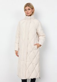 Lange, gewatteerde pufferjas in lichtbeige met een hoge kraag, zijzakken en een rechte snit. Bevat een ruitpatroon en een ritssluiting.