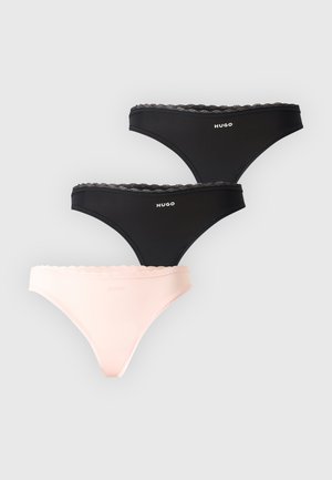 Femme aux longs cheveux foncés portant un soutien-gorge de sport noir marqué Hugo et un sous-vêtement noir assorti, debout avec une main sur la hanche.