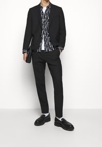 Completo nero con rever a punta, abbinato a una camicia bianca con un motivo astratto in bianco e nero. Completato con mocassini neri.