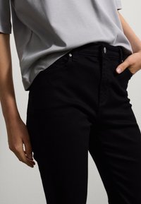 Jeans neri in cotone con taglio slim, tasche frontali e cuciture a vista. Indossati con una maglietta a maniche corte grigia chiara.