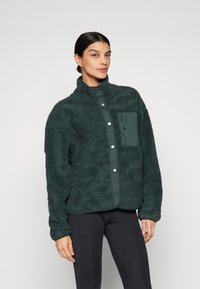 Fleecejacke in Dunkelgrün mit weicher Textur, hohem Kragen, Druckknopfverschluss und einer Reißverschlusstasche auf der linken Brust.